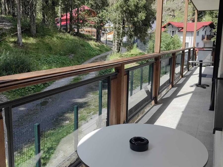 En balkon med et bord og stole, der ud til en flod og et skovområde med bygninger i baggrunden.