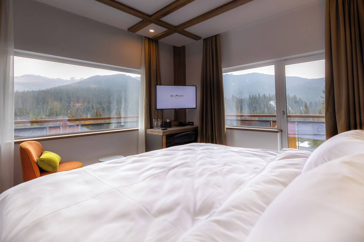 A hotel room with a large bed, a desk with a computer, a TV on the wall, and a view of mountains through large windows.