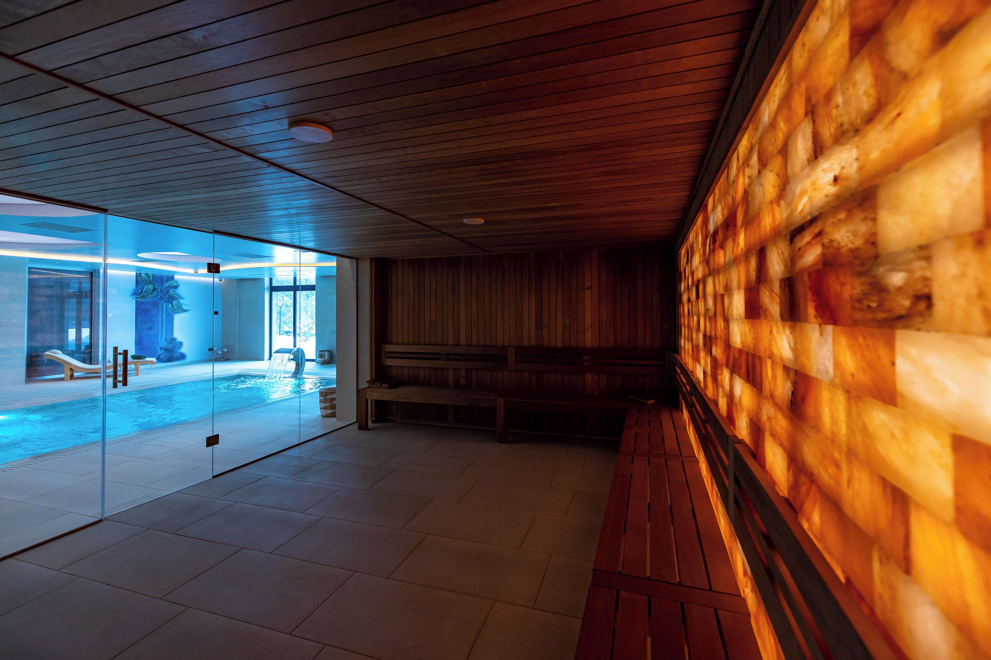 Un espace piscine intérieur moderne avec des murs en verre, des bancs en bois et un sol carrelé.