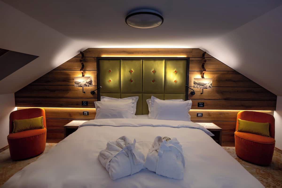 A modern hotel room with a sloped ceiling, a large bed with white linens, two red armchairs, and a wooden headboard with wall-mounted lights.