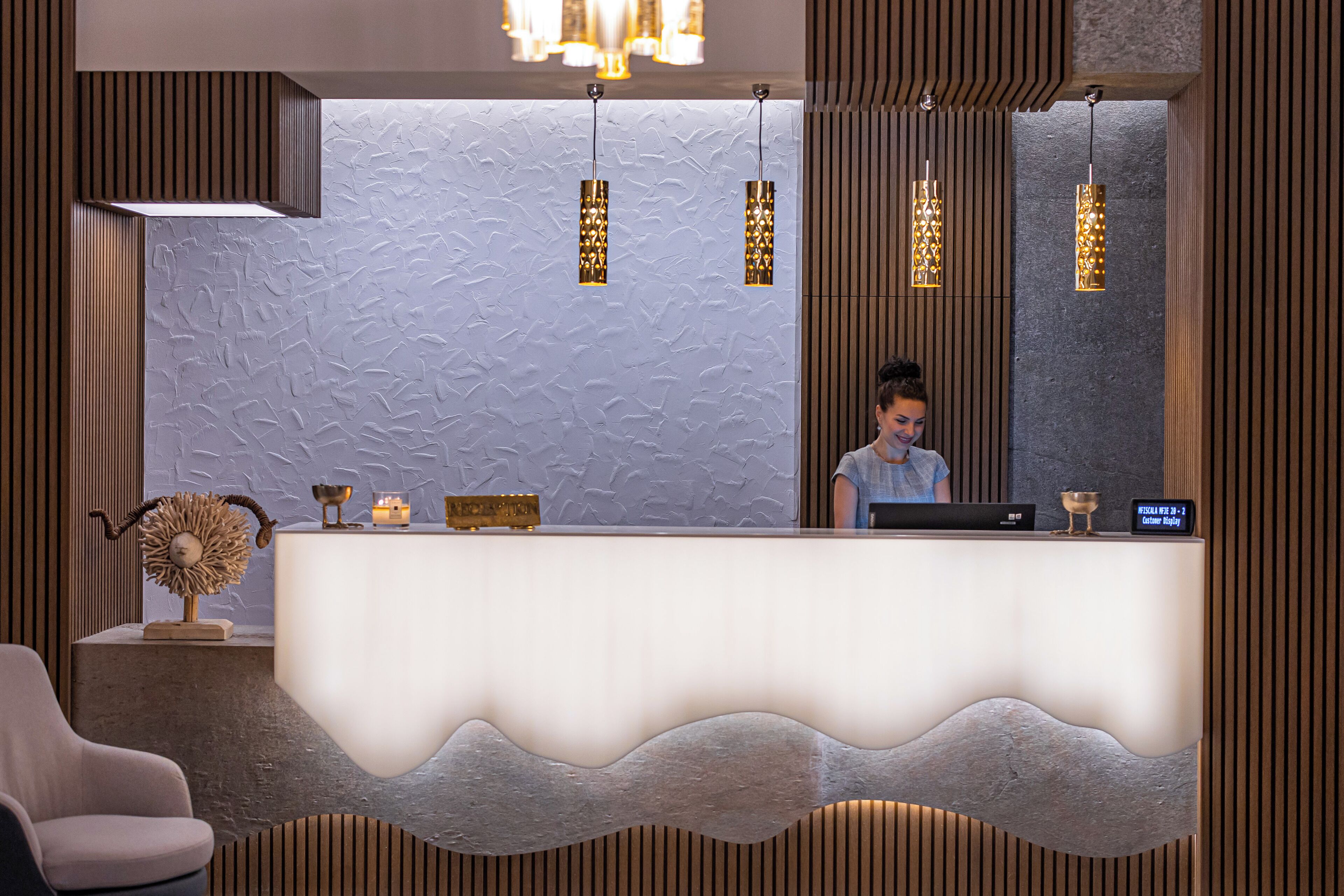 A reception area with a modern design, featuring a textured wall, a reception desk with a wave-like front, and pendant lighting.