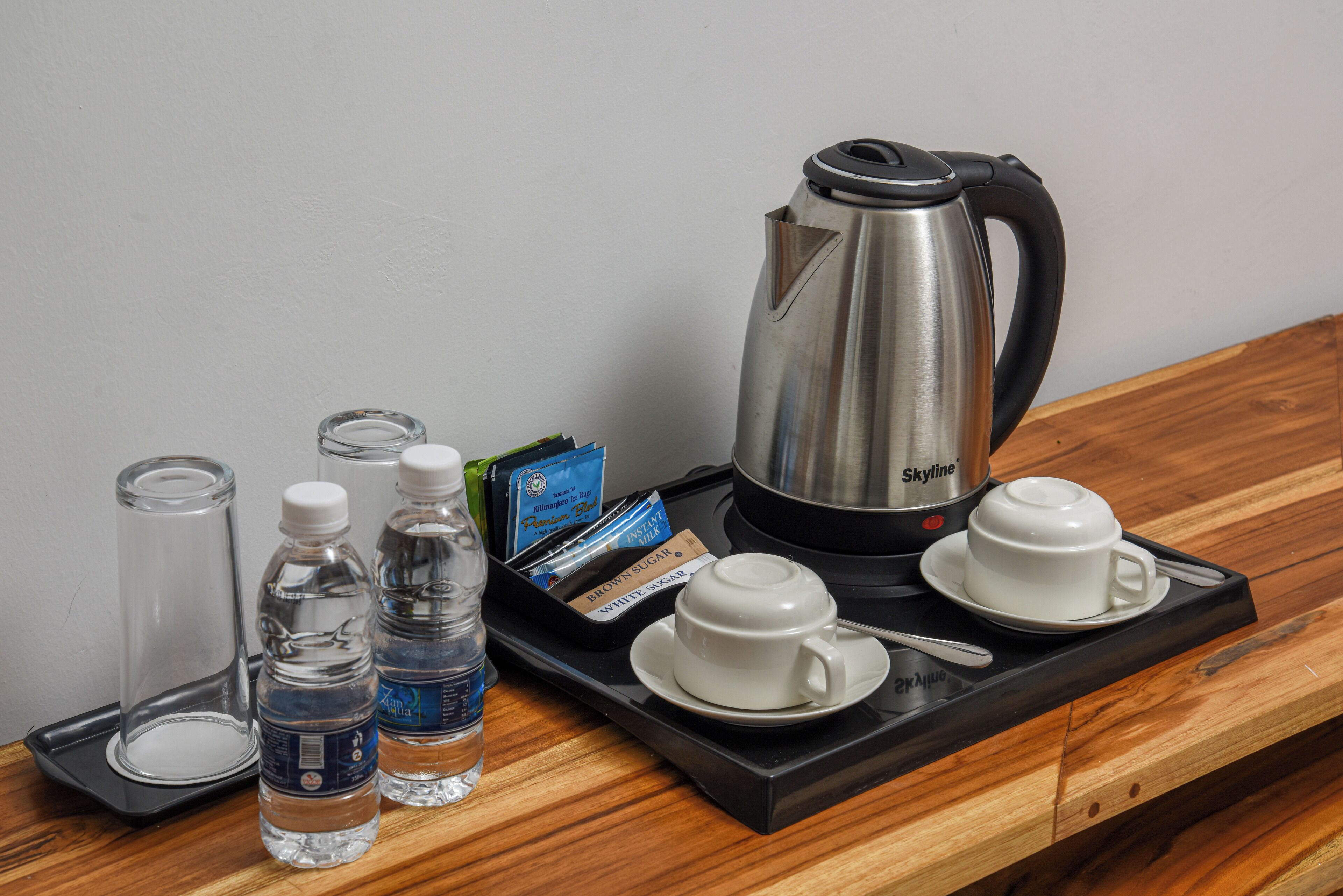 A tray with a stainless steel kettle, two white cups, a spoon, and three water bottles on a wooden table.