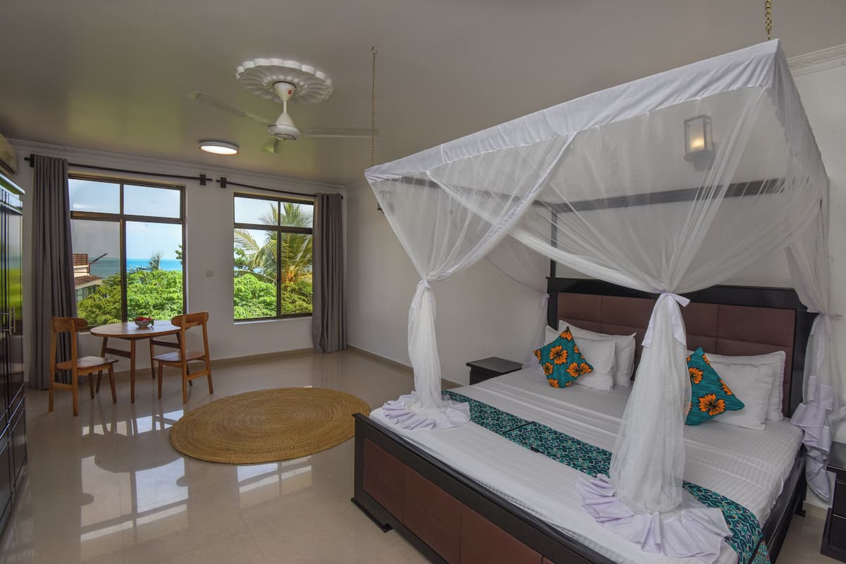 Un dormitorio con cama con dosel, una zona de comedor con mesa y sillas, y ventanas amplias con vistas al verdor.