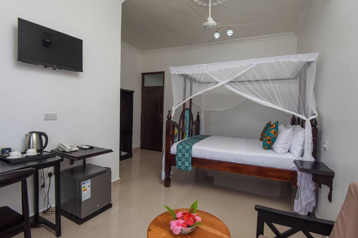 Habitación de hotel con cama, escritorio, refrigerador pequeño, televisor y mesita de noche con lámpara.