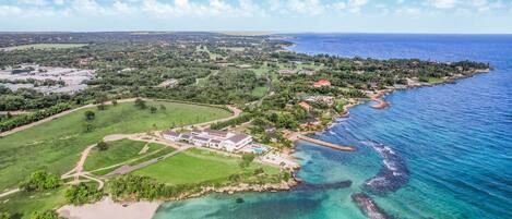 Vista aérea de un complejo turístico costero con un edificio grande, una playa y aguas azules cristalinas.