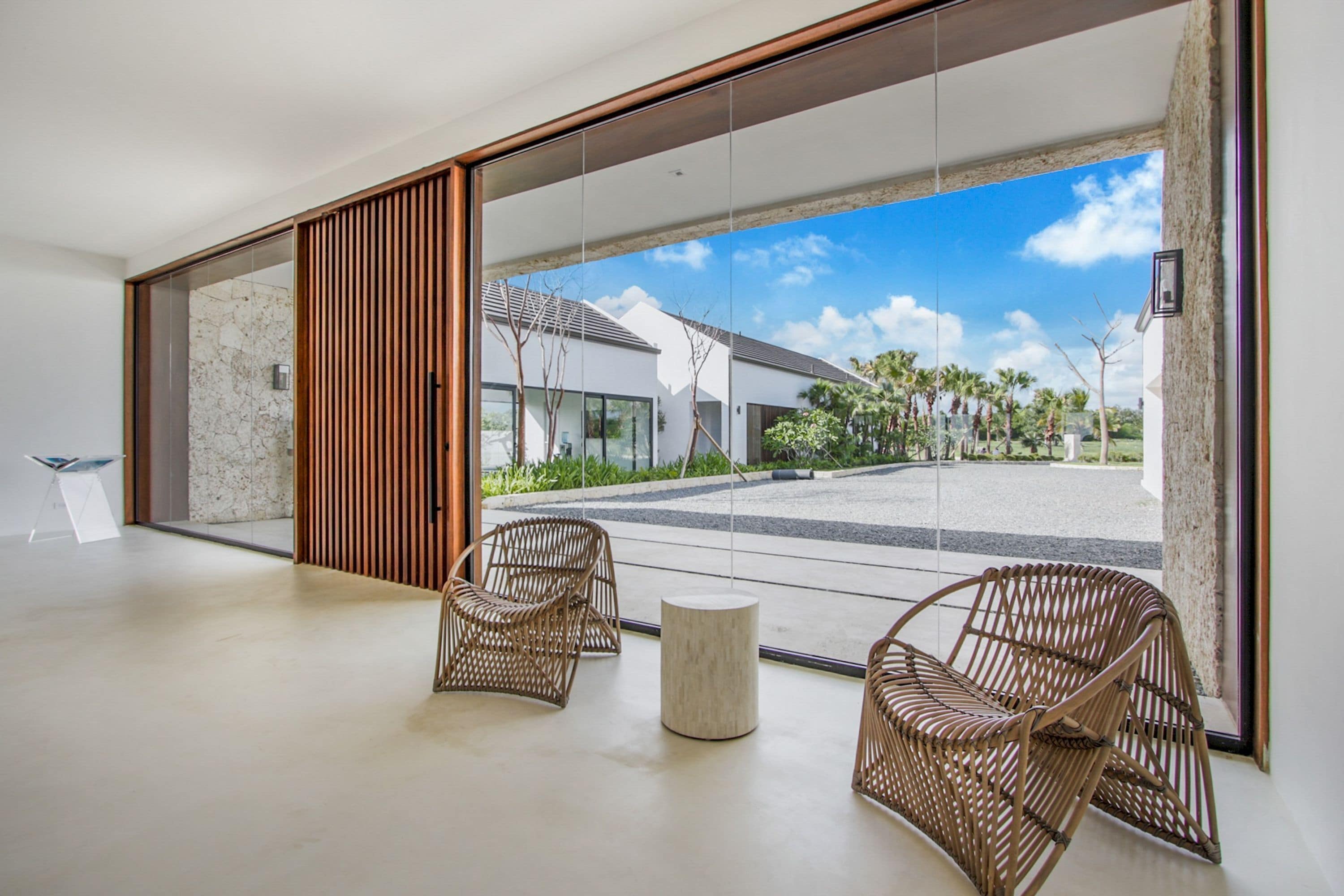 Un interior moderno con sillas de mimbre y una mesa redonda, puertas de cristal amplias que dan a un patio con vistas a un edificio y palmeras.
