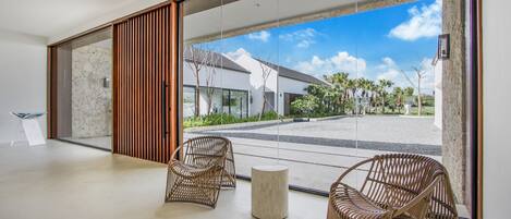 Un interior moderno con sillas de mimbre y una mesa redonda, puertas de cristal amplias que dan a un patio con vistas a un edificio y palmeras.