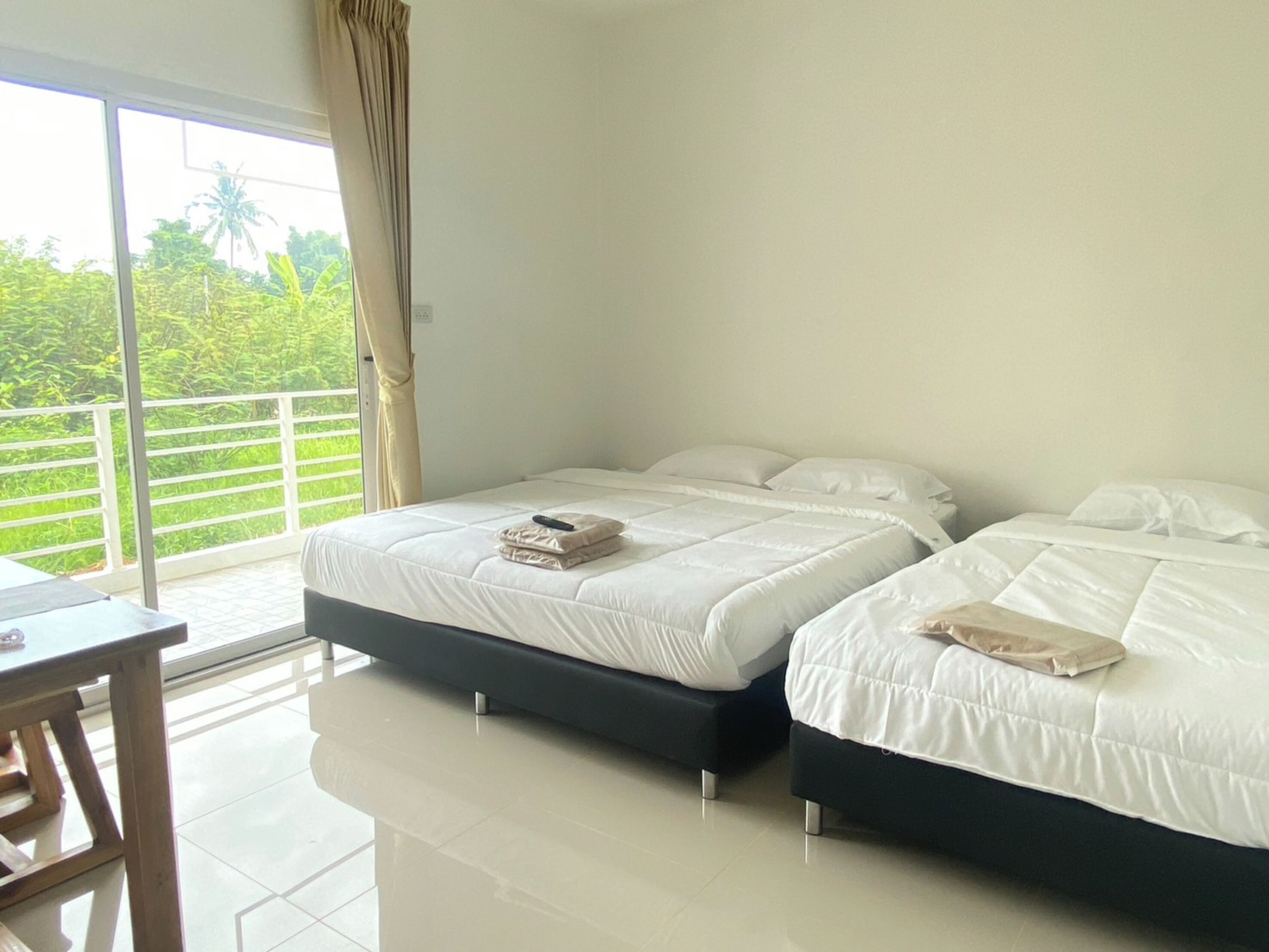 A hotel room with two beds, a balcony with a view of greenery, and a table.