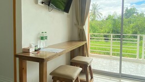 A room with a wooden desk, two stools, a TV mounted on the wall, and a view of greenery through a sliding glass door. - Dino Homestay (Mae Taeng)