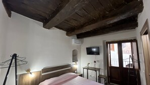 Een slaapkamer met een houten plafond, een bed met een roze dekbedovertrek, een nachtkastje, een kleine tafel met een lamp, een aan de muur bevestigde televisie en een raam met uitzicht op het omliggende landschap.