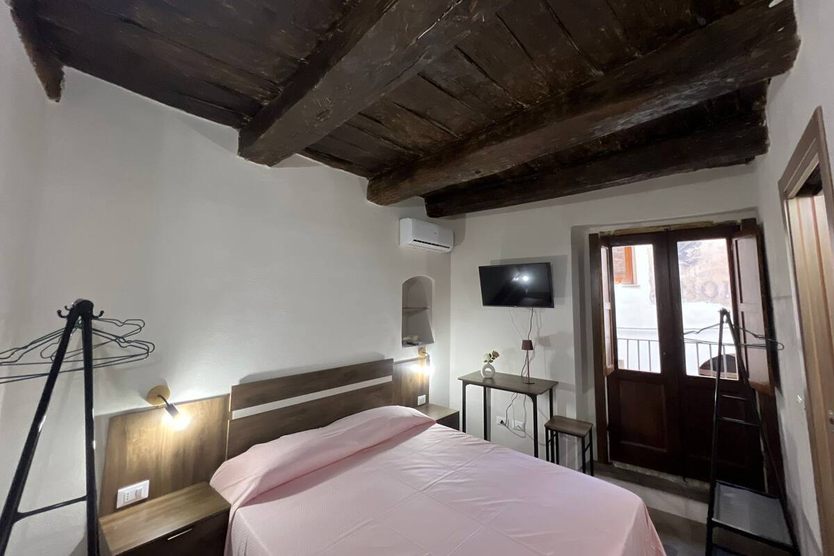 Un dormitorio con techo de madera, una cama con colcha rosa, una mesita de noche, una pequeña mesa con una lámpara, un televisor montado en la pared y una ventana con vistas al exterior.