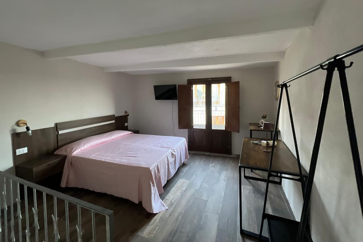 Un dormitorio con cama, escritorio, silla, televisor y ventana con contraventanas abiertas.