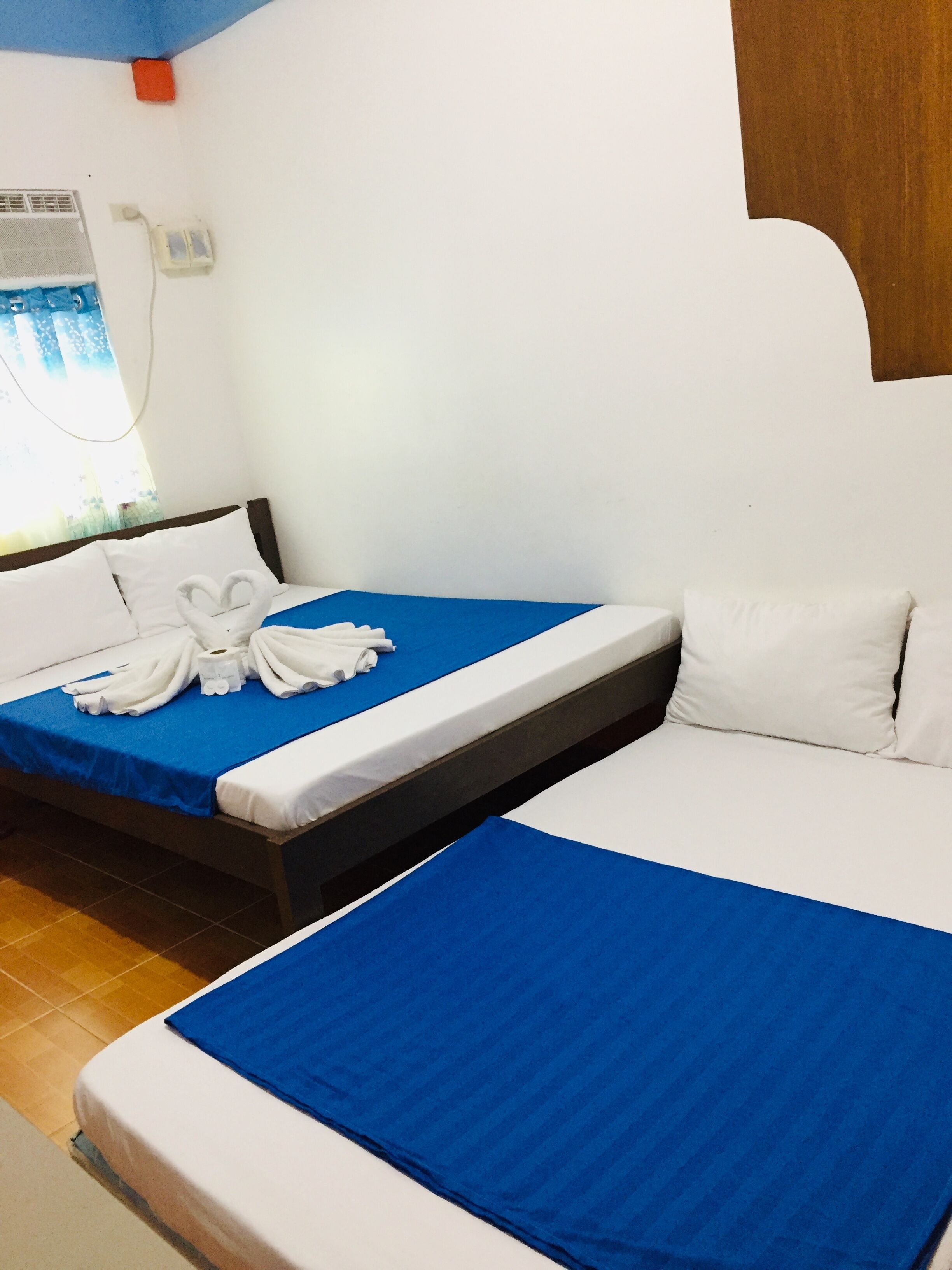 A hotel room with two beds, each with a blue bedspread and white pillows. There is a window with a view of greenery outside.