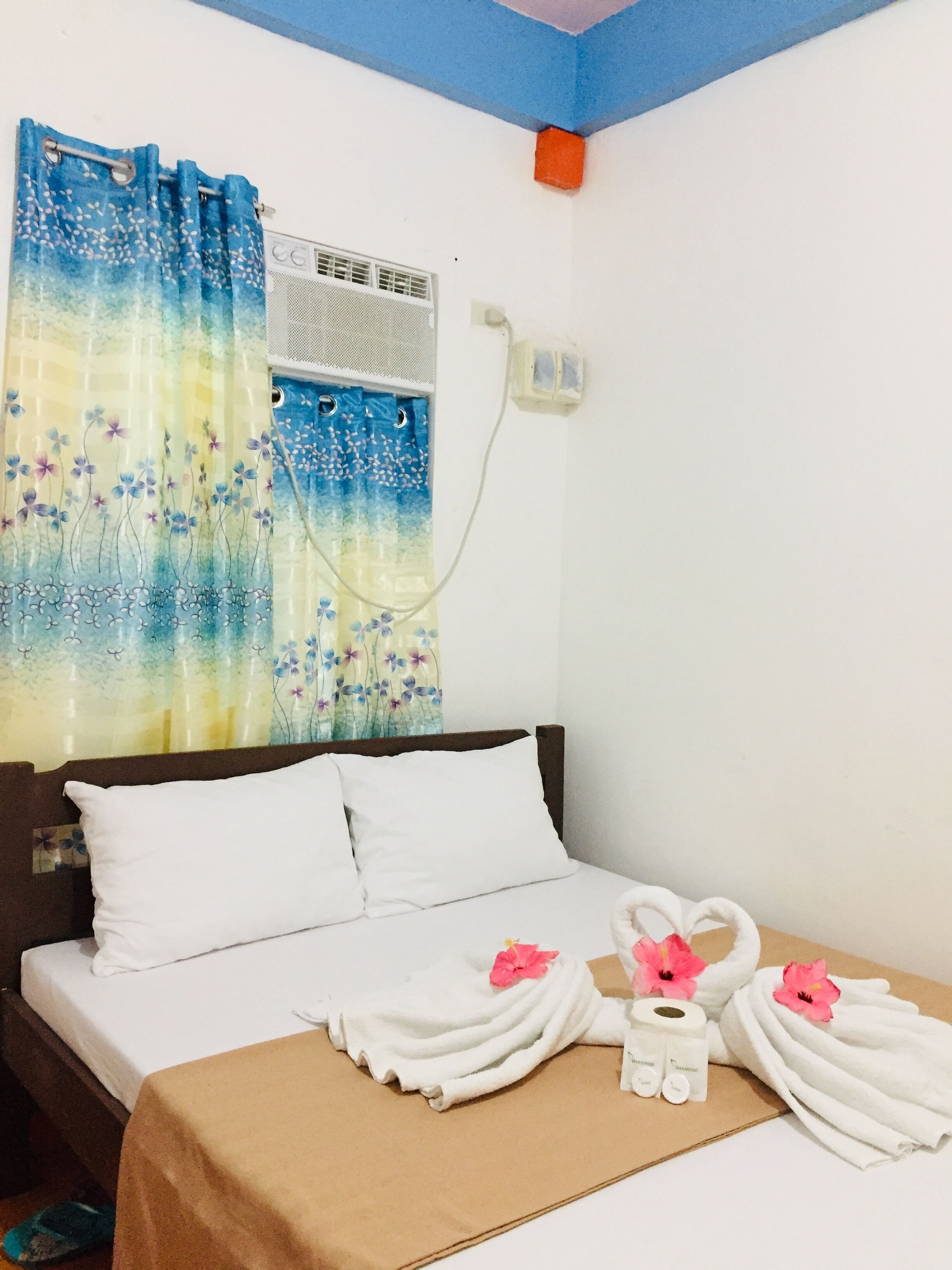 A hotel room with a bed, white pillows, a brown bedspread, a floral curtain, a wall-mounted air conditioner, and a towel rack with towels and toiletries.