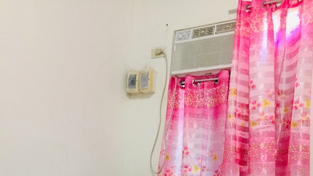 Ein Hotelzimmer mit Bett, weißen Kissen, einem schwarzen Kopfteil, einer weißen Wand, einem Fenster mit rosa blumigen Vorhängen und einer Wand-Klimaanlage.