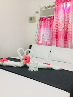 A hotel room with a bed, white pillows, a black headboard, a white wall, a window with pink floral curtains, and a wall-mounted air conditioning unit. - Bebs and Jocel Apartment (Panglao)