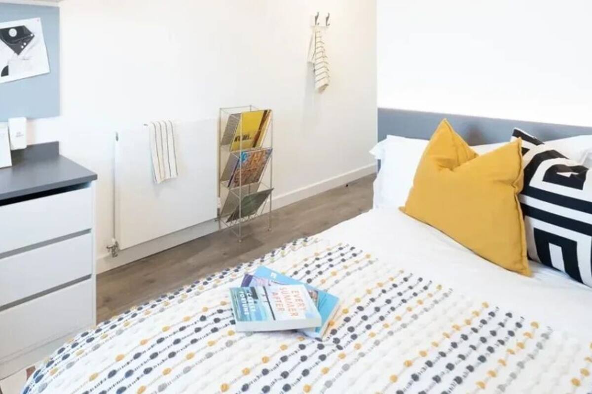 A bedroom with a bed, a desk, a chair, a radiator, and a shelf with books and a vase of flowers.