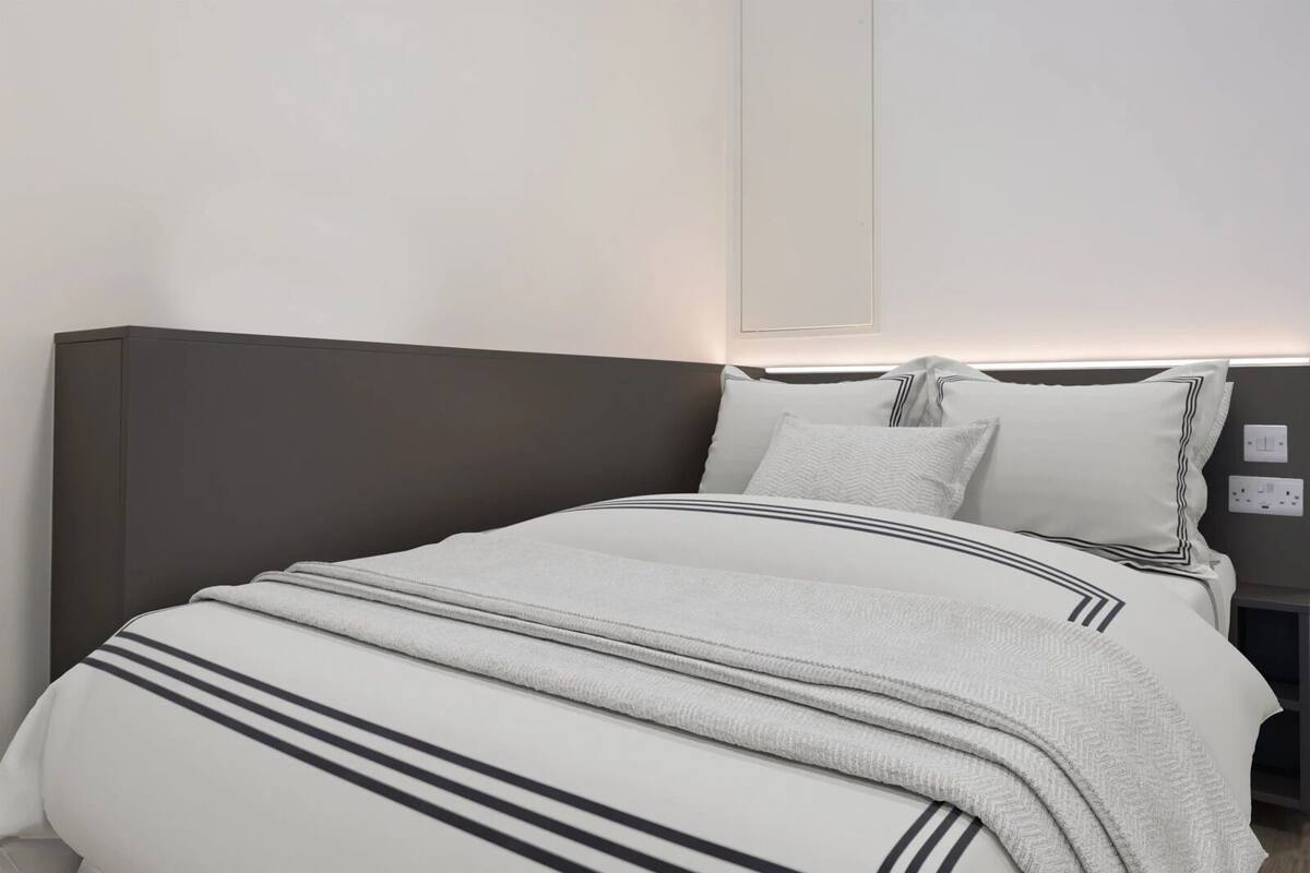 A neatly made bed with a striped blanket and pillows, a bedside table with a lamp, and a wall-mounted light fixture.