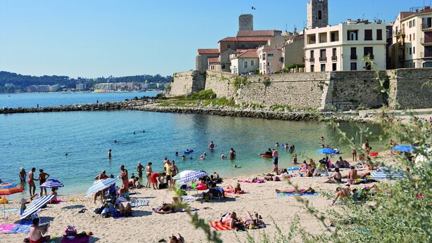 Une plage où l’on voit des gens nager et bronzer, un mur de pierre et des bâtiments dotés de balcons.
