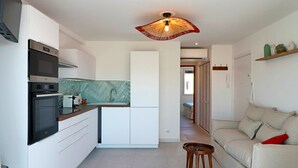 Une cuisine compacte dotée d’appareils électroménagers modernes, un coin repas avec une table et des chaises, et un salon avec un canapé et des coussins.
