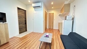 A compact living space with a wooden floor, a small table, a sofa, a TV, a kitchen area, and an air conditioning unit. - SOLASIA residence Kitaueno501 (Tokyo)