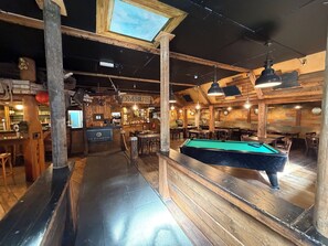 A billiards table, wooden bar counter, and seating area in a rustic-themed venue.