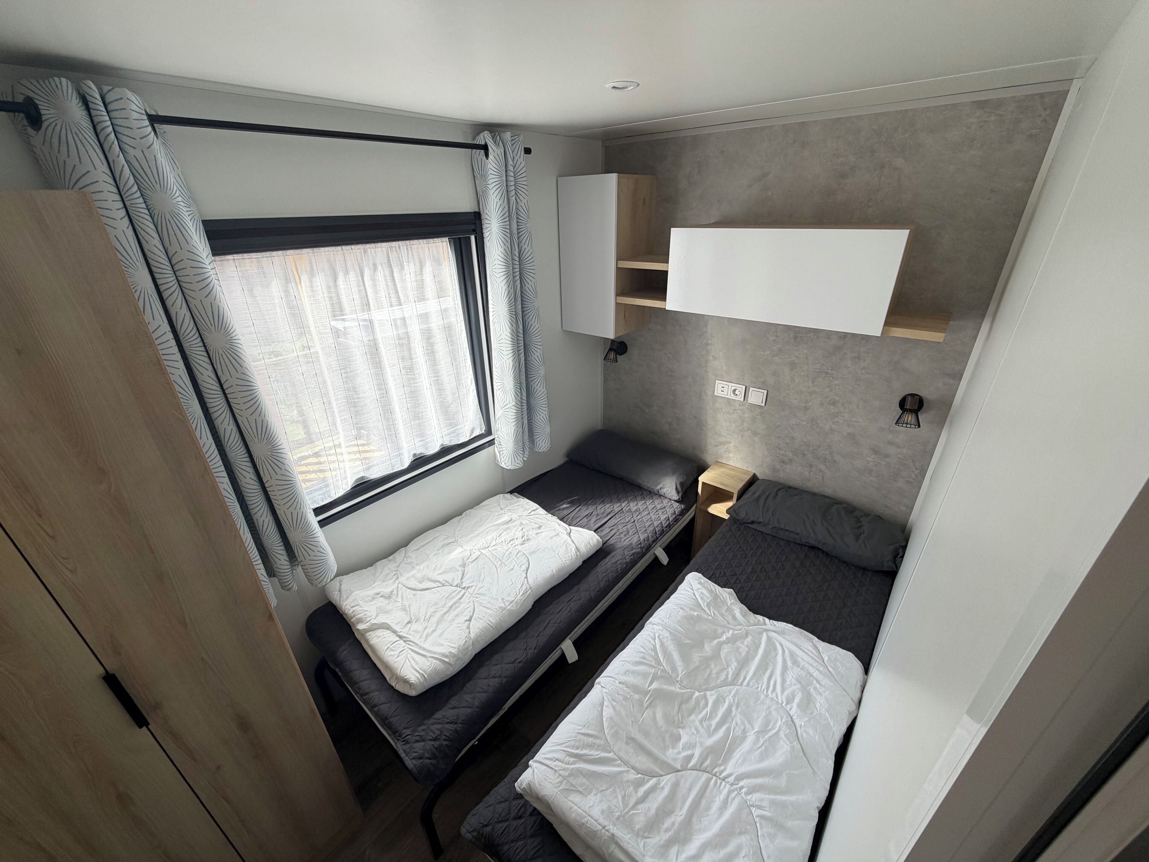 Habitación pequeña con dos camas, una ventana con cortinas, un estante y una lámpara fijada a la pared.