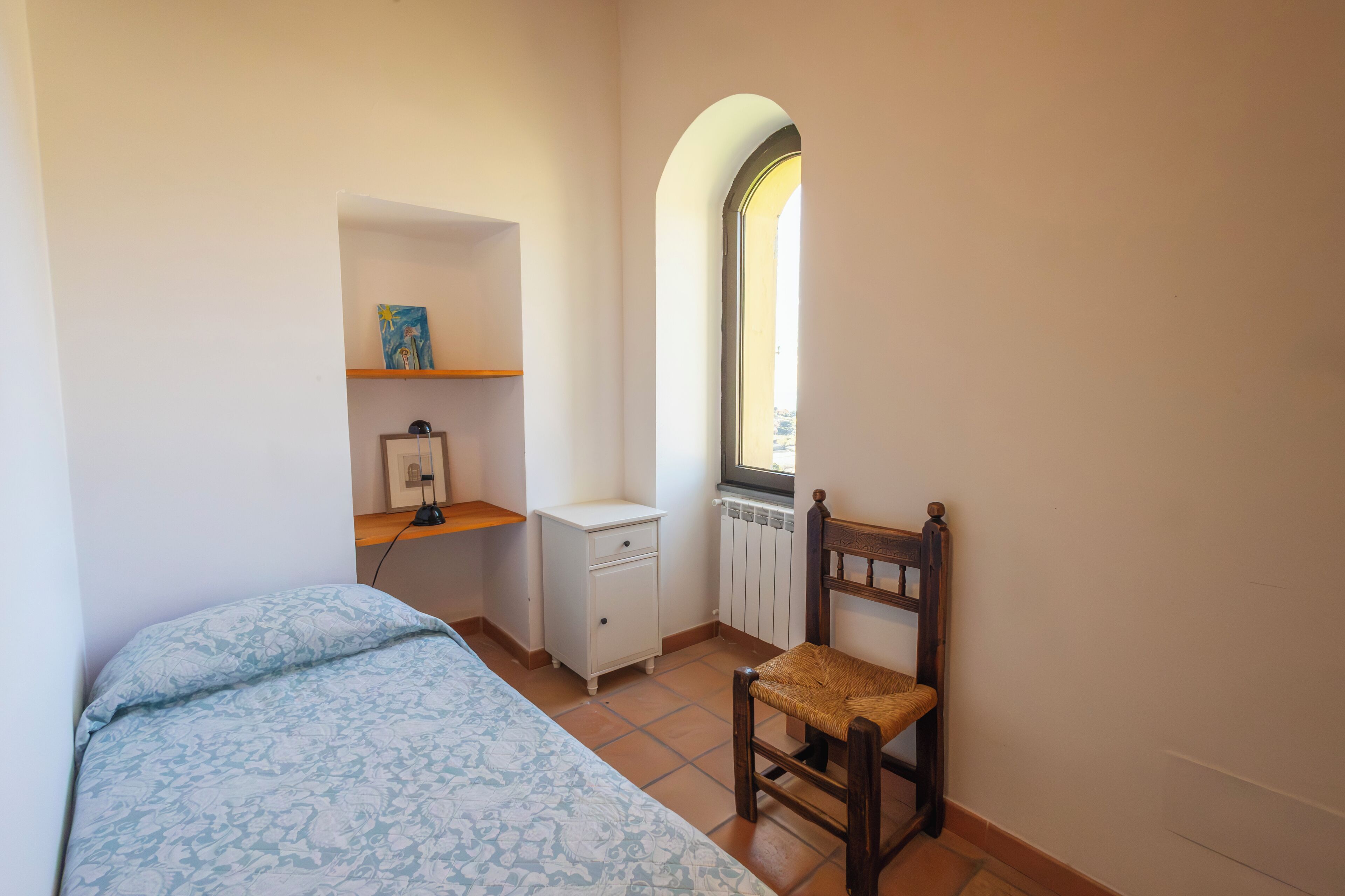 Un dormitorio con cama, una silla, un pequeño tocador blanco, una estantería con un cuadro y un ventanal arqueado.