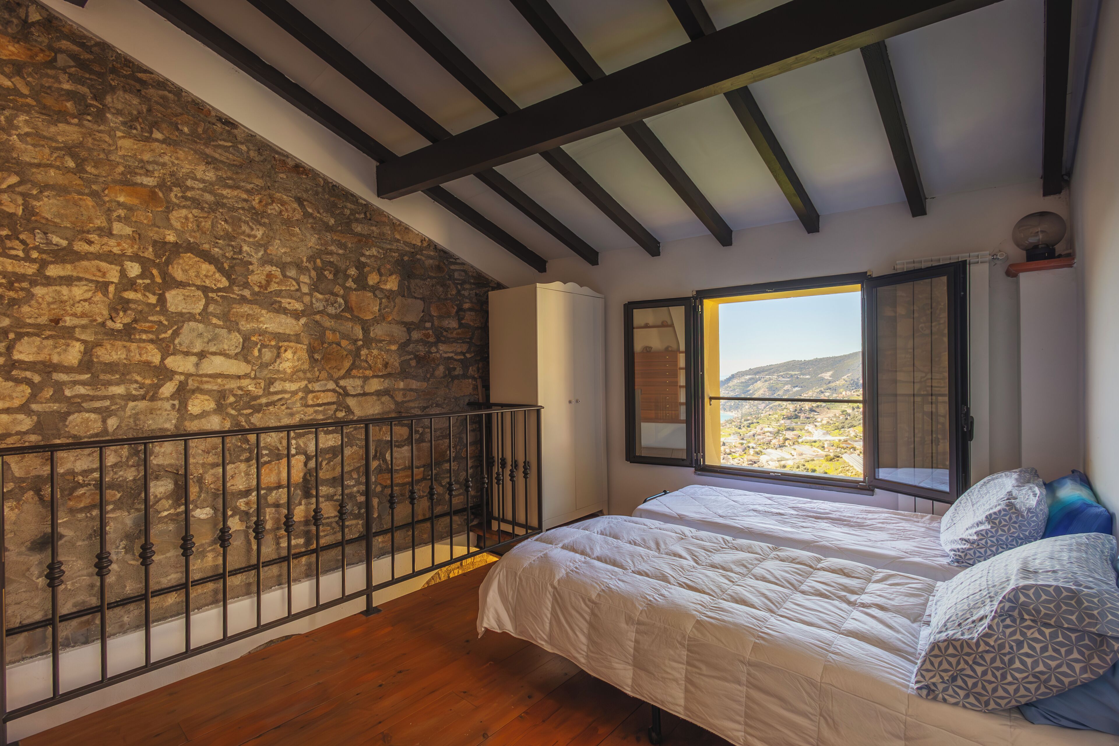 A bedroom with a stone wall, wooden floor, a bed with white bedding, and a window with an open shutter.