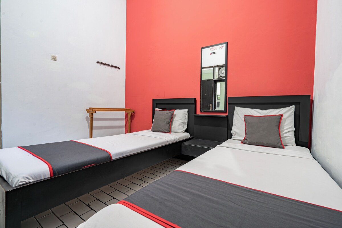 Une chambre d’hôtel avec deux lits simples, une chaise en bois, un miroir et un mur d’accent rouge.