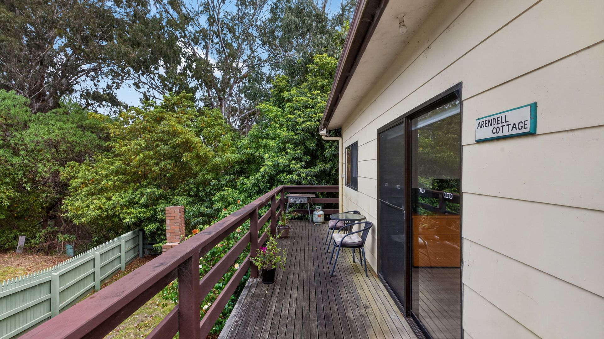 A wooden deck with a glass door leading to a room, surrounded by lush greenery and a sign indicating 'ARRENDELLE COTTAGE'.