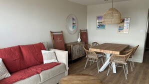 Un salon chaleureux avec une table à manger en bois, des chaises en osier, un canapé gris agrémenté de coussins rouges et une table basse ronde.