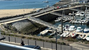 Un port de plaisance sur la côte, avec des bateaux amarrés, une route menant à la plage et un phare visible au loin.