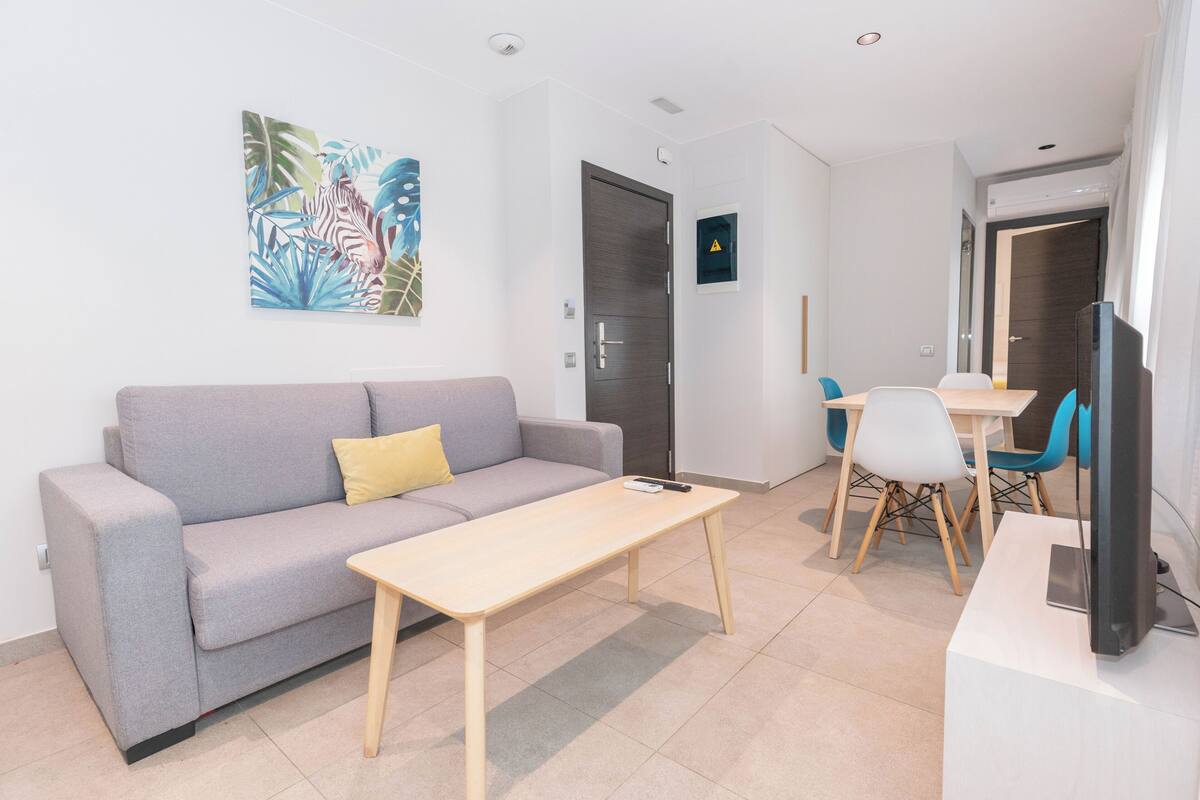 Un salon moderne avec un canapé gris, une table basse en bois, un téléviseur et un coin repas avec des chaises.