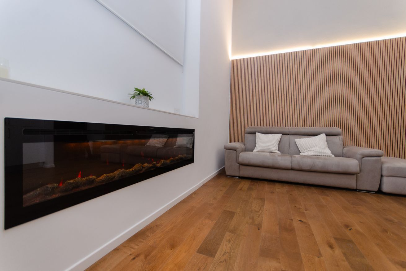 A modern living room with a large flat-screen TV mounted on the wall, a grey sofa with white cushions, and a wooden floor.