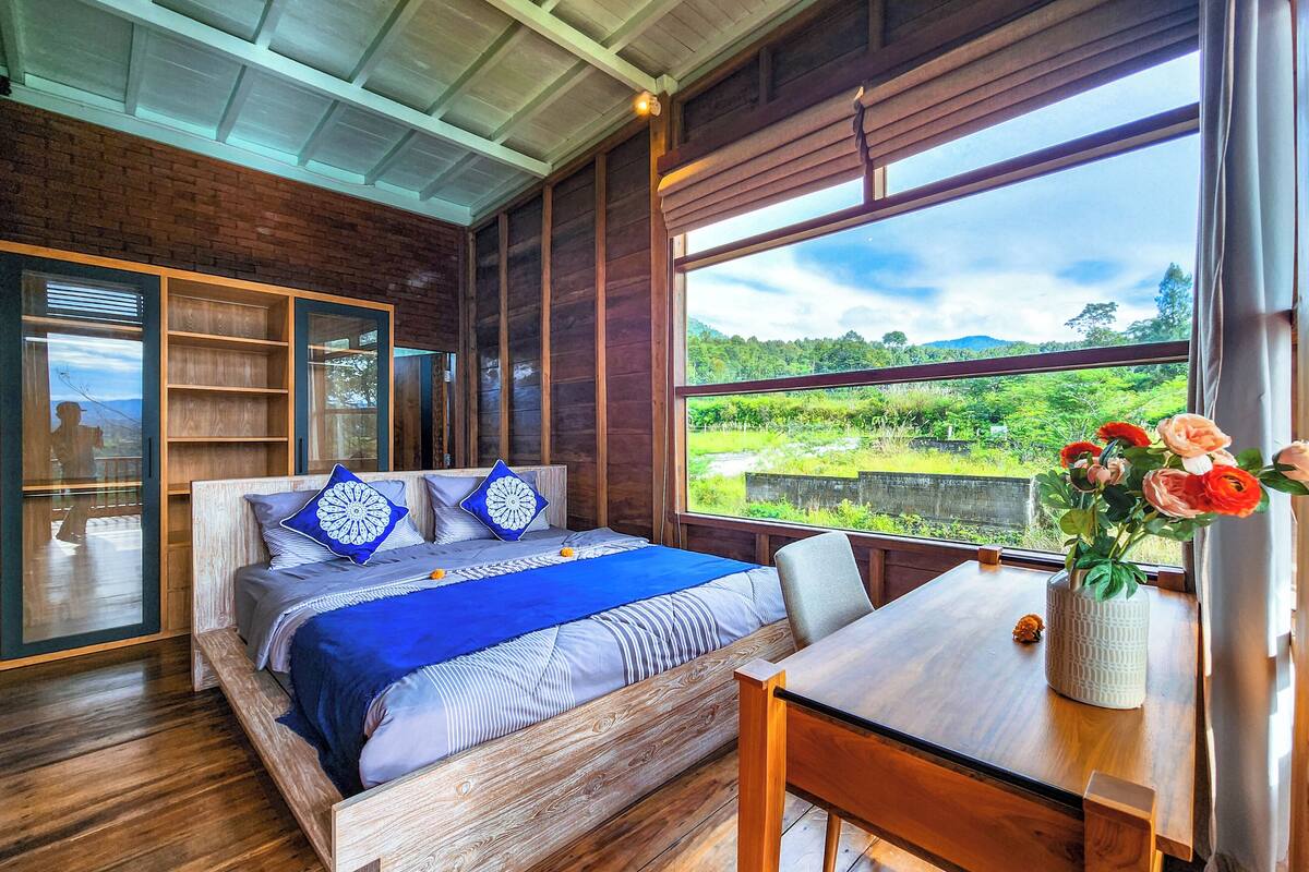 A bedroom with a wooden bed, a view of greenery, and a table with a vase of flowers.