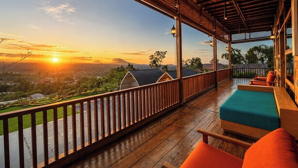 A wooden deck with a view of a sunset over a landscape, a wooden railing, and a seating area with cushions. - Mountain Healing Villa by Nagisa Bali (Baturiti)