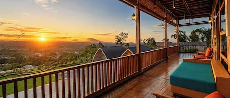 Una terraza de madera con vistas a una puesta de sol sobre el paisaje, barandilla de madera y un área de descanso con cojines.