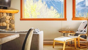 A cozy room with a stone fireplace, a large window with a mountain view, a comfortable sofa, a wooden chair, and a small table with glasses. - Ha Ling Haven - Penthouse with Shared Rooftop Hot Tub (Canmore)