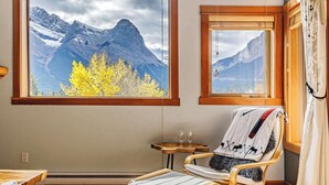 A room with large windows offering mountain views, a wooden rocking chair, a small table with a glass, and a curtain. - Ha Ling Haven - Penthouse with Shared Rooftop Hot Tub (Canmore)