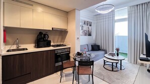 Eine kompakte Küche mit Gasherd, Mikrowelle und Kaffeemaschine. Ein Essbereich mit kleinem Tisch und zwei Stühlen. Das Zimmer verfügt über ein großes Fenster mit Vorhängen und Blick ins Freie.