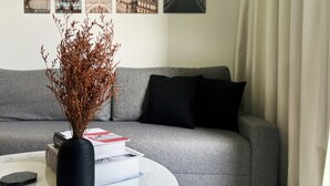 Ein Wohnzimmer mit einem grauen Sofa, einem weißen Couchtisch, einer schwarzen Vase mit Trockenpflanzen und einem Stapel Bücher.