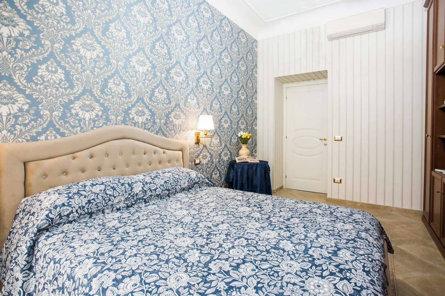 A bedroom with a tufted headboard, a patterned bedspread, a bedside table with a lamp, a vase of flowers, and a white door.