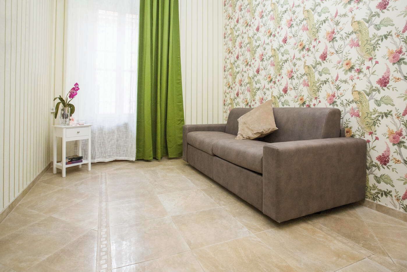 Un salon avec un canapé gris, une petite table d’appoint blanche, un rideau vert et un papier peint à motifs floraux.
