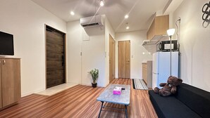 A modern, well-lit room with wooden flooring, a small table with a box, a potted plant, a teddy bear, and a TV on the left wall.