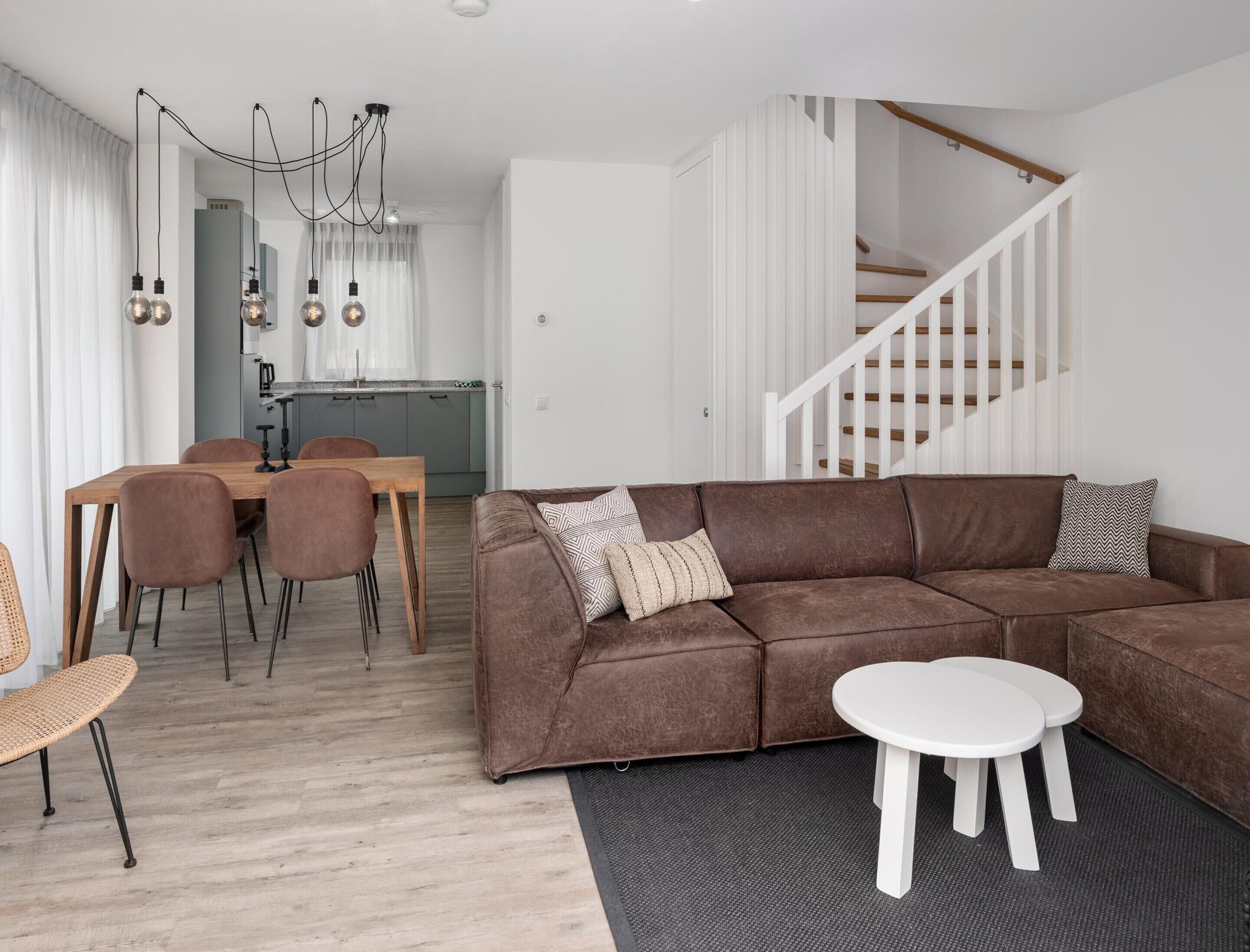 A modern living room with a brown sectional sofa, a dining table with chairs, and a staircase.
