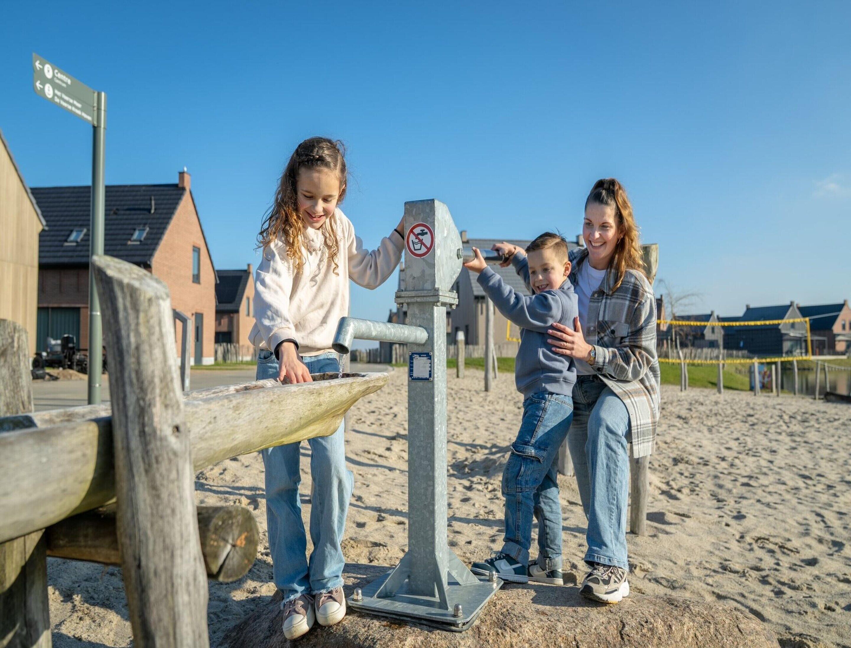 Una famiglia interagisce con una pompa per l'acqua su una spiaggia sabbiosa, con edifici sullo sfondo.