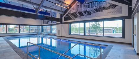 Ein Hallenbad mit klarem, blauem Wasser, umgeben von gefliesten Wänden und einer hohen Decke mit Metallbalken.