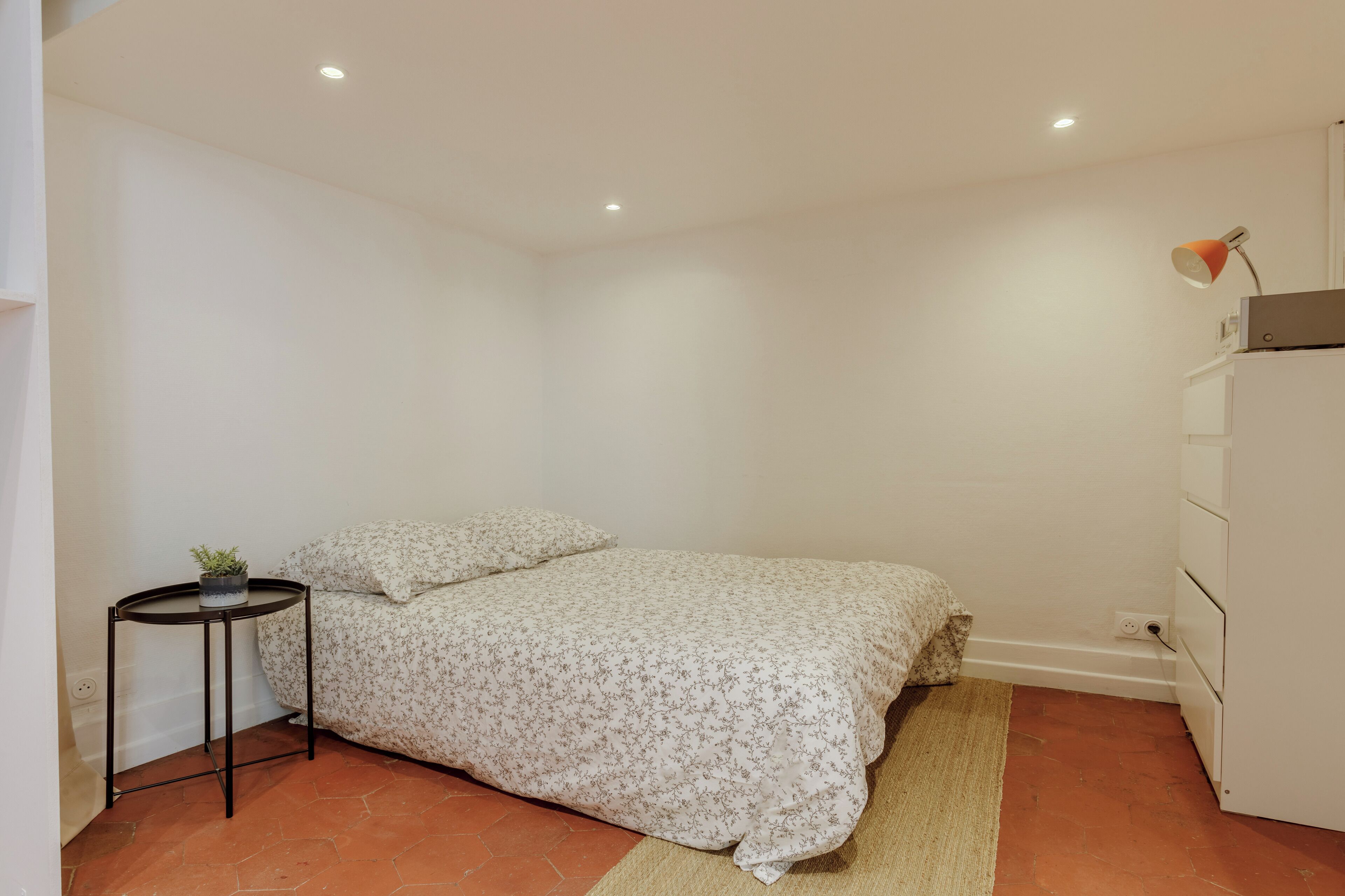 A bedroom with a bed, a small side table with a plant, a lamp, and a dresser.