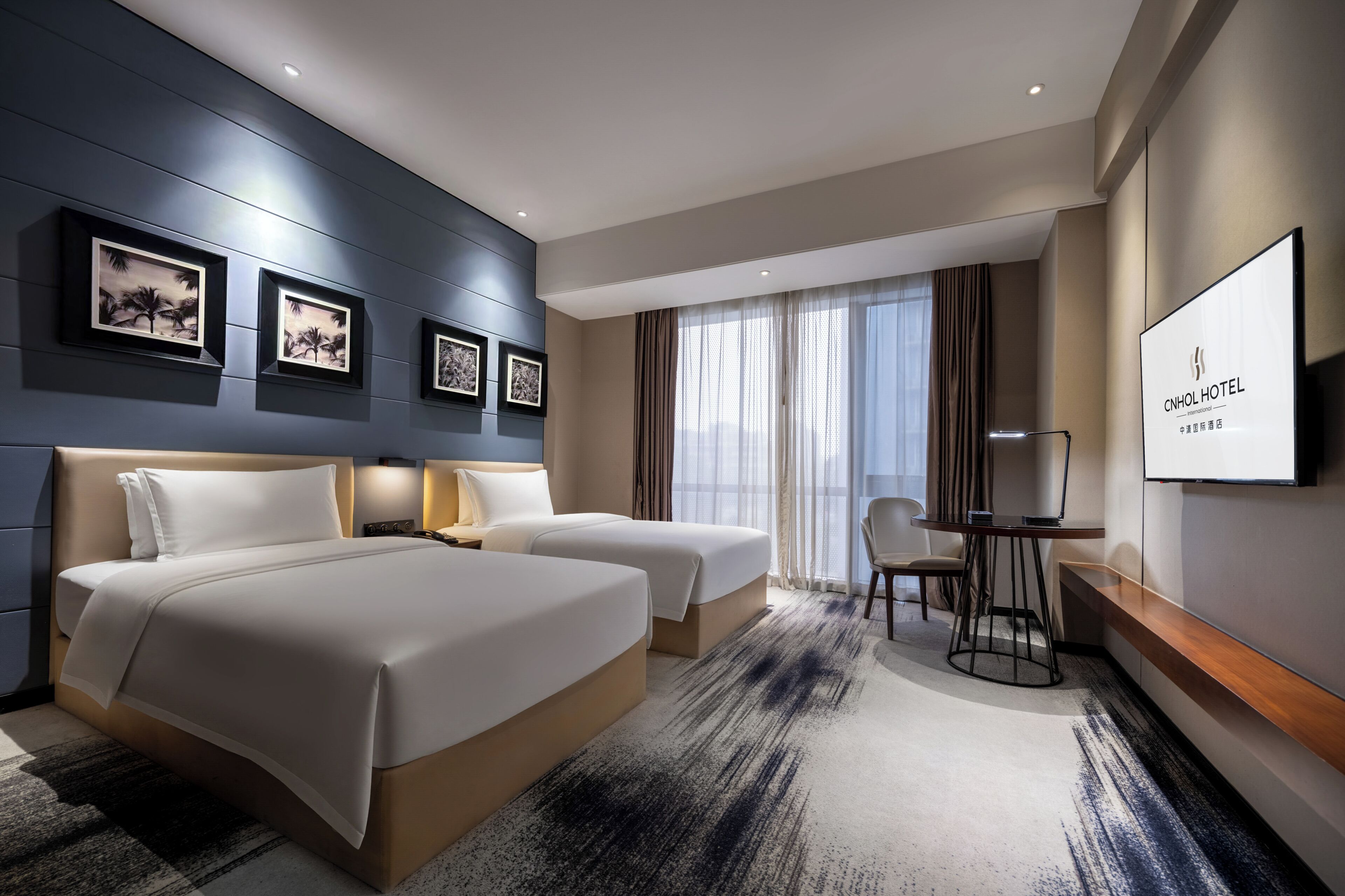A hotel room with two beds, a desk, a chair, a TV, and framed pictures on the wall.